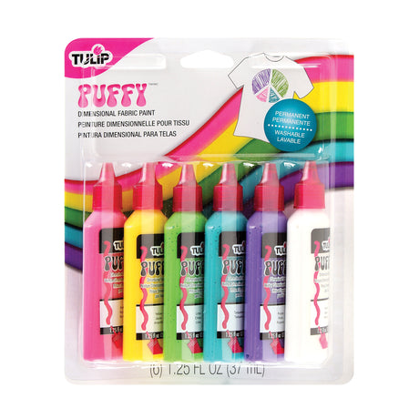 Tulip Puffy Dimensional Fabric Paint packaging, a blister pack displaying six upright squeeze bottles with red nozzle caps, in pink, yellow, lime, turquoise, purple and white. The card backing features a rainbow stripe design, a white t-shirt graphic decorated with coloured sections, and a blue “Permanent, Washable” badge.