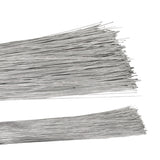 Two bundles of straight silver florist wire laid horizontally against a white background, the fine metallic strands fanning out at the cut ends. The long, slender wires have a bright silvery-grey finish and lie in loose overlapping layers, a few crossing diagonally across the stacks.