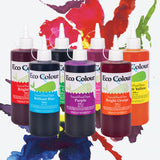A group of Eco Colour Liquid Food Dye bottles in an assortment of colours, each with a white nozzle cap and whale illustration label. Clearly visible labels include Brilliant Blue, Purple, and Bright Orange, with additional bottles partially visible behind, set against colourful watercolour paint splashes.