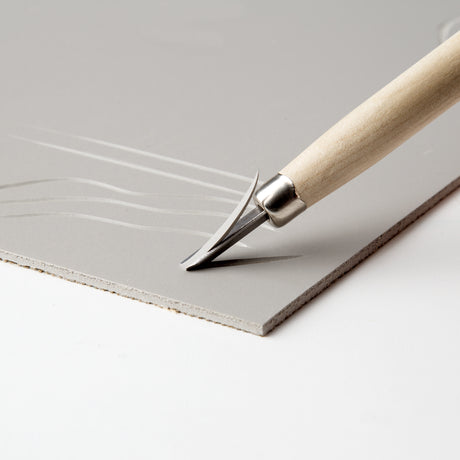Close-up photograph of a lino carving tool cutting into a grey Silk Cut Lino Tile. The sharp blade lifts a thin curl of lino as smooth curved lines are carved into the surface, demonstrating controlled mark making and precision cutting.