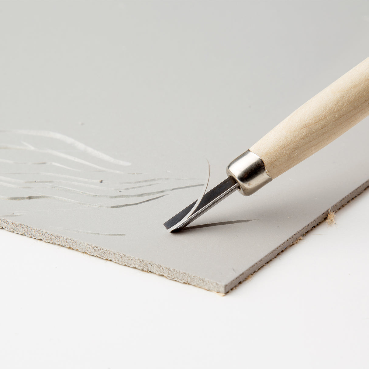 Close-up of a wooden-handled carving gouge cutting into a Lino Tile Smooth Carve surface, creating clean, precise lines. The metal blade lifts a delicate curl from the pale grey linoleum, demonstrating the smooth carving surface. The tile's layered edge is visible.