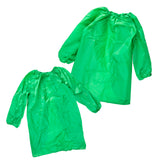 Two green Zart smocks laid out together, a smaller one in front and a larger one behind, showing the size difference between them. Both share the same elasticated round neckline, long sleeves with elasticated cuffs, and a curved, uneven hemline dipping lower at the centre front.