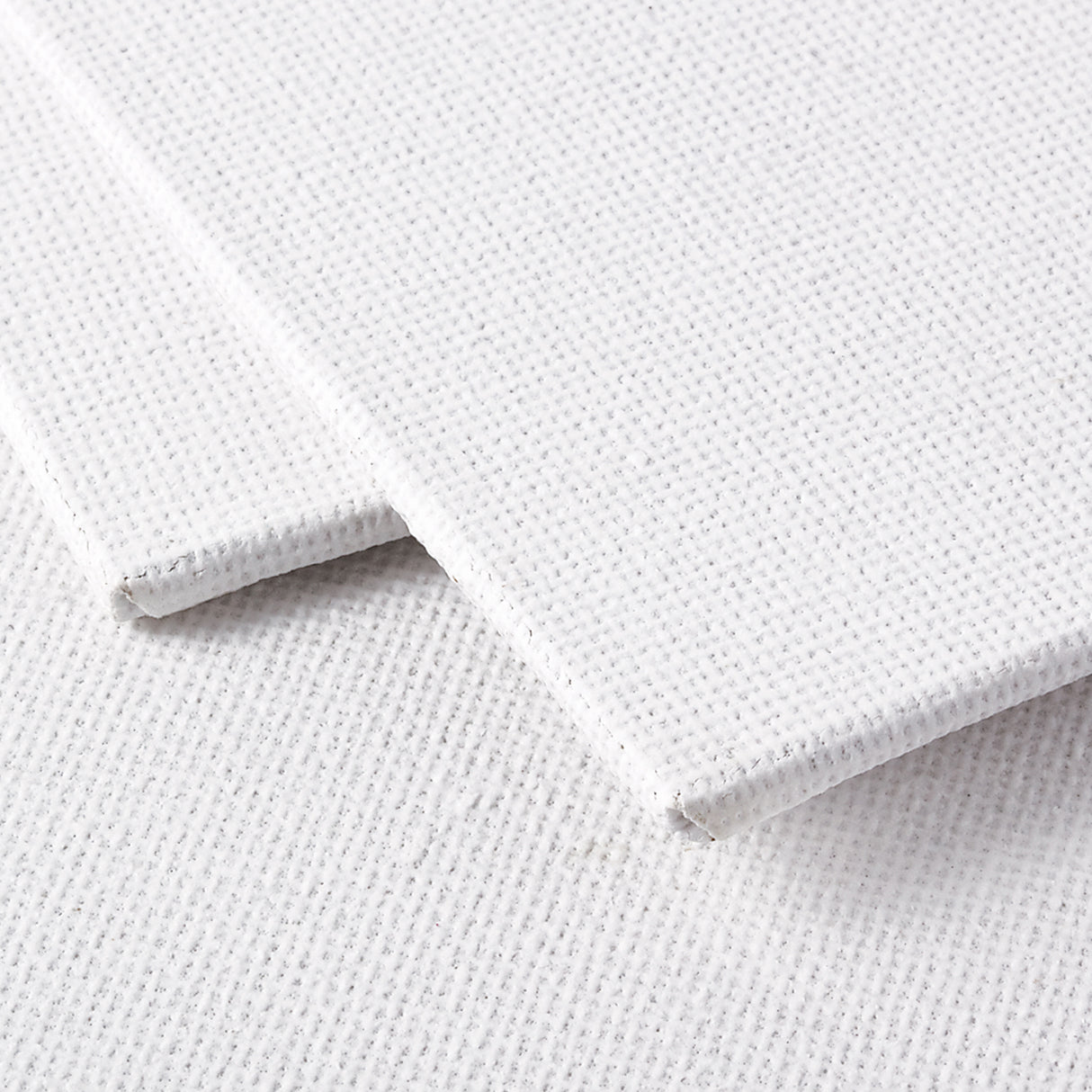 Close-up view of two white canvas boards stacked slightly offset. The woven canvas texture is clearly visible across the surface, with the top board lifted to reveal the firm board backing and clean wrapped edges. The surface shows a fine grid weave typical of primed canvas used for painting.