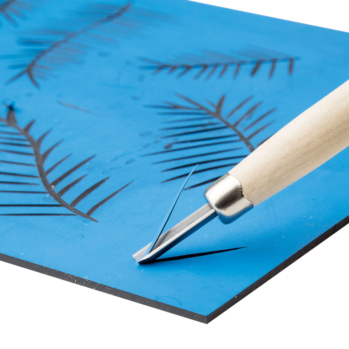 Close-up of a carving tool cutting into a blue Velvet Print Lino Tile. The blade lifts a thin curl of material, revealing smooth carved lines forming leaf patterns across the surface, demonstrating the tile’s soft, easy-to-carve texture.