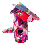 A standing dragon-like creature made from Zart Acetate Heavyweight Sheets, its body covered in overlapping scale-shaped pieces coloured in pink, purple, blue, red and gold with speckled metallic detailing. A drawn eye sits on the red face, showing a three-dimensional sculptural application.