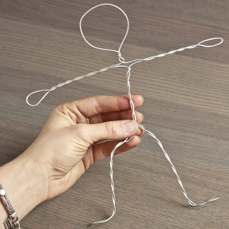 Hand holding pliable Armature Wire twisted into a simple stick-figure shape, demonstrating flexibility and ease of bending. The silver wire is partially twisted to form arms, legs and a looped head, shown against a wooden tabletop background.