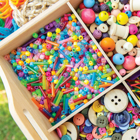 A close-up of Zart Basics plastic beads filling one compartment of a wooden tray, showing translucent and opaque pony beads and coloured tube beads in a vivid rainbow mix. Adjacent compartments hold wooden beads, spools and assorted buttons, with braided cord resting at the top left.