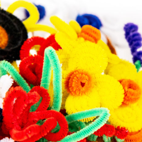 A close-up of Zart Basics chenille stems bent and coiled into loops and spirals, demonstrating how easily they shape. Fuzzy stems in yellow, orange, red, mint green, blue, purple, black and white twist together, some wound into tight coils against a soft white background.