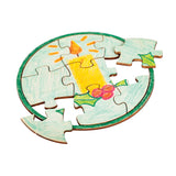A round jigsaw sample made using a Zart Mini Jigsaw Round puzzle, hand-coloured with a festive candle design in yellow and orange, surrounded by holly leaves and berries in green and red, on a pale blue background with a green painted border. Two pieces sit detached.