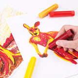 A hand holds a red Ochre Slick, actively colouring the body of an outlined kangaroo drawing in warm red and pink tones. A capped yellow stick lies below, with another yellow and red stick pair resting above. A partly coloured tree and sunburst landscape sits at left.