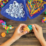 A child's hands pressing Joggle Eyes onto a foam clay creature at a wooden table, with a partitioned blue tray of loose Joggle Eyes and coloured craft sticks nearby, plus buttons in the top corner. Completed foam clay figures with Joggle Eyes sit to the left and right.