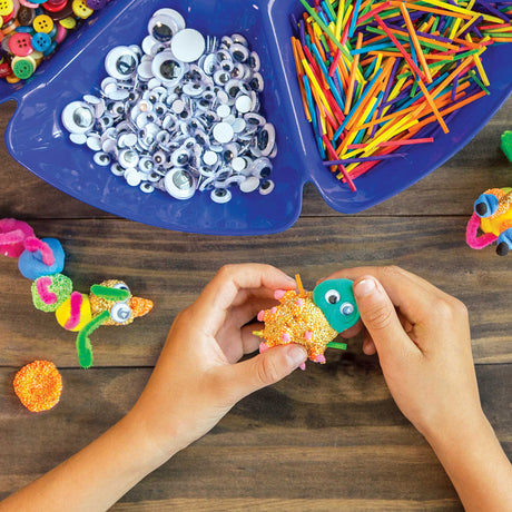 A child's hands pressing Joggle Eyes onto a foam clay creature at a wooden table, with a partitioned blue tray of loose Joggle Eyes and coloured craft sticks nearby, plus buttons in the top corner. Completed foam clay figures with Joggle Eyes sit to the left and right.