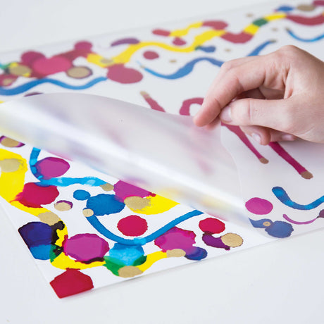 A hand lifts a translucent Zart Laminating Pouch over a colourful abstract artwork featuring bright pink, blue, yellow and gold paint marks. The frosted pouch surface softly blurs the artwork beneath while showing the flexible plastic texture and smooth protective finish.