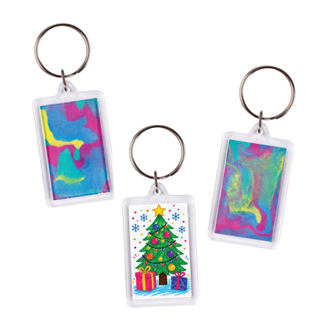 Three Zart Rectangle Key Ring Tags shown as sample artworks with decorative inserts. The left tag displays a swirling blue, pink, and yellow abstract colour pattern. The centre tag features a colourful illustrated Christmas tree with gifts. The right tag shows a blended pink, blue, and green swirl design.