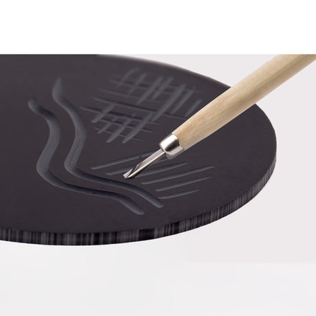 Close-up of a Mastercut Print Lino Round block being carved with a V-shaped lino cutting tool. The dark grey lino surface shows carved grooves, curved lines and textured marks. The tool’s wooden handle and metal blade are positioned mid-cut, demonstrating carving detail.