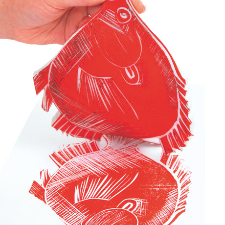 Hand lifting a carved fish-shaped soft cut block coated in red ink. Below, a red print impression of the fish appears on white paper. Fine carved lines create crosshatching and contour detail across the body and fins.