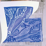 A sheet of white paper is peeled back to reveal a blue print made from a foam plate, showing a bird perched on a branch. Beneath the paper, the inked foam surface shows indented lines forming leaves, feathers and branch details. The print sits on newspaper during the printing process.
