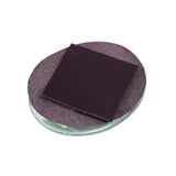 A single Zart Adhesive Magnet square, matte black, resting on top of a round glass disc lined with brown felt backing. One corner of the magnet is peeled slightly upward, revealing the felt beneath and showing the flat, thin profile of the square against the circular base.