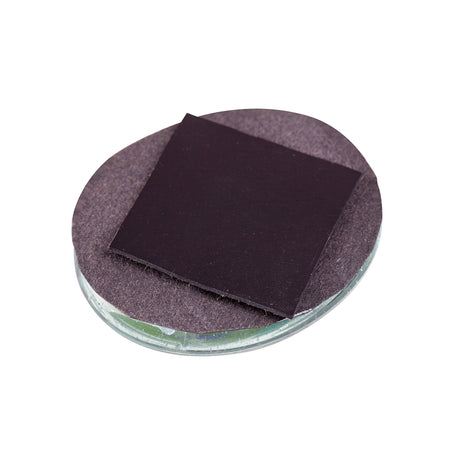 A single Zart Adhesive Magnet square, matte black, resting on top of a round glass disc lined with brown felt backing. One corner of the magnet is peeled slightly upward, revealing the felt beneath and showing the flat, thin profile of the square against the circular base.