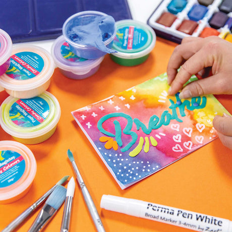 Hands use Zart Paper Magiclay Candy Colours to decorate a painted card. Open tubs of clay sit nearby with brushes and markers, showing mixed media use for lettering and surface decoration.