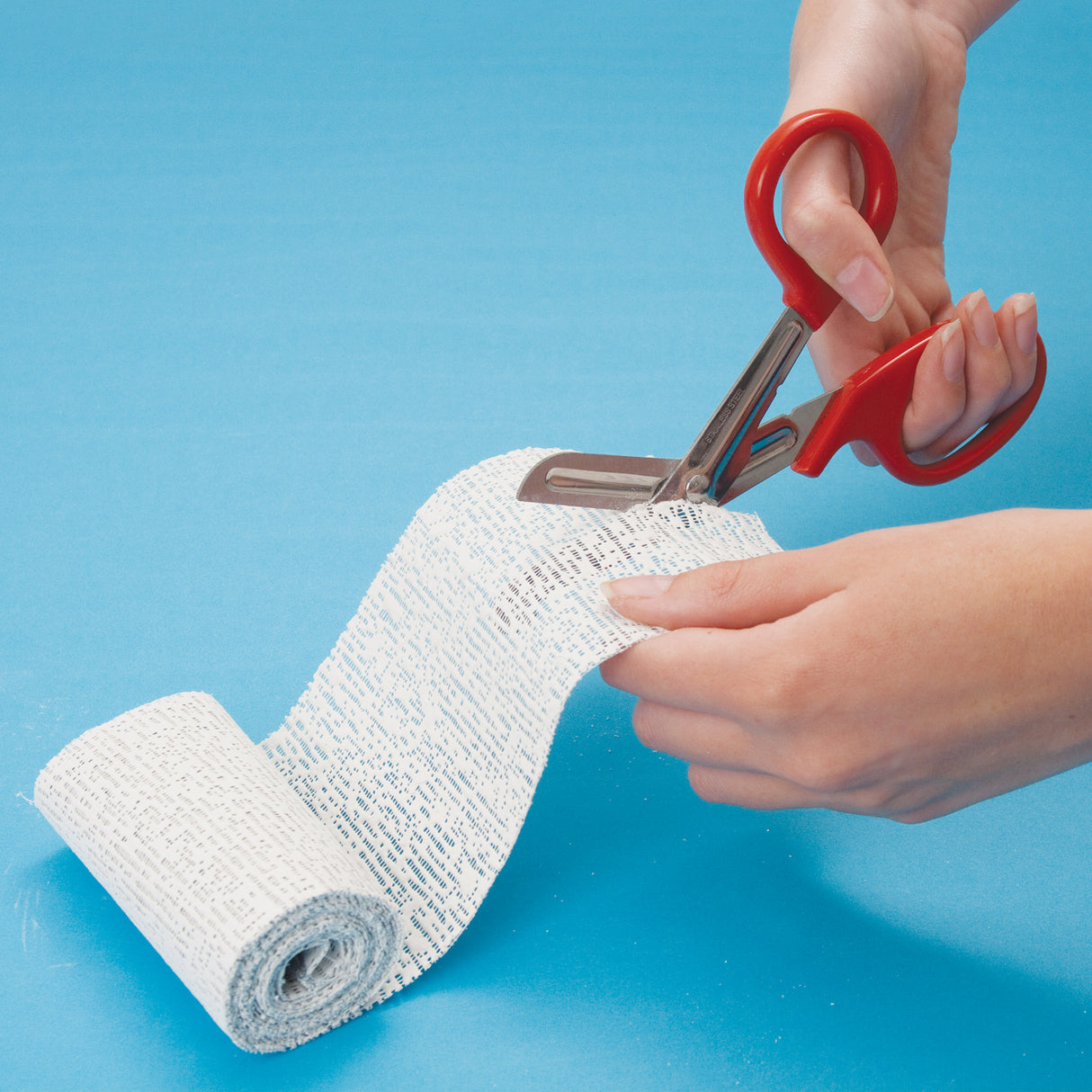 Hands using red-handled scissors to cut a strip from a roll of plaster-impregnated cloth on a blue surface. Demonstrates how the material can be easily cut to size. No text annotations shown.
