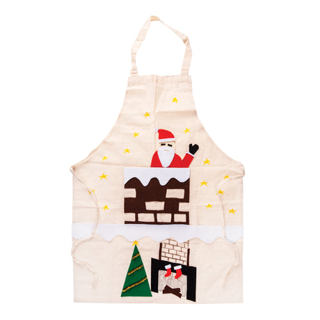 A Zart Calico Apron decorated as a festive Christmas scene. A red and white Santa Claus figure waves from behind a brown chimney with a snowy top. Gold stars scatter across the natural fabric, alongside a green tree with gold diagonal stripes and a fireplace holding two red stockings.