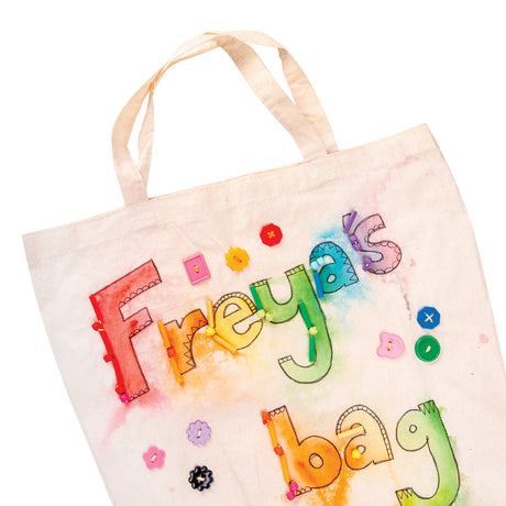 A close-up of a Zart Calico Bag with Handles decorated as a sample artwork, with "Freyja's bag" hand-painted in rainbow bubble letters outlined in black, each letter threaded with colourful beads, and scattered flower and geometric buttons stitched across the surrounding fabric.