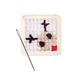 A small square panel of woven white and cream yarn mounted on a light wooden frame with punched holes around its edges. Dark purple X-shaped and oval stitches sit across the surface, with a red patch in one corner. A silver needle rests alongside.