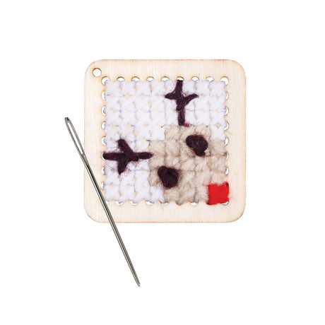 A small square panel of woven white and cream yarn mounted on a light wooden frame with punched holes around its edges. Dark purple X-shaped and oval stitches sit across the surface, with a red patch in one corner. A silver needle rests alongside.