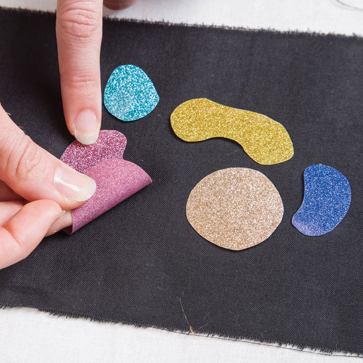 A hand peels a pink Zart Glitter Iron-On Sheet cut-out from its backing onto black fabric, alongside teal, gold, tan and blue glitter pieces cut into organic, leaf-like shapes. Demonstrates the sheet's soft, pliable texture and vibrant colour when applied to dark fabric.