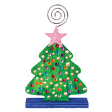 A decorated Zart Photo Holder Tree, painted green with a scattering of coloured gem and pearl embellishments across the tree shape and a glittery pink star topper. The tree stands on a blue-painted rectangular base, with the silver spiral wire clip rising above the star.