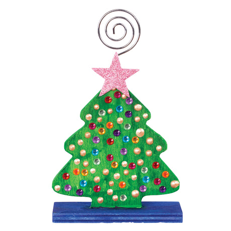 A decorated Zart Photo Holder Tree, painted green with a scattering of coloured gem and pearl embellishments across the tree shape and a glittery pink star topper. The tree stands on a blue-painted rectangular base, with the silver spiral wire clip rising above the star.
