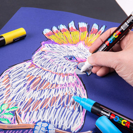 Hand using a white POSCA paint marker to add details to a colourful cockatoo drawing on dark blue paper. Other POSCA markers sit nearby, showing their bright, opaque lines.