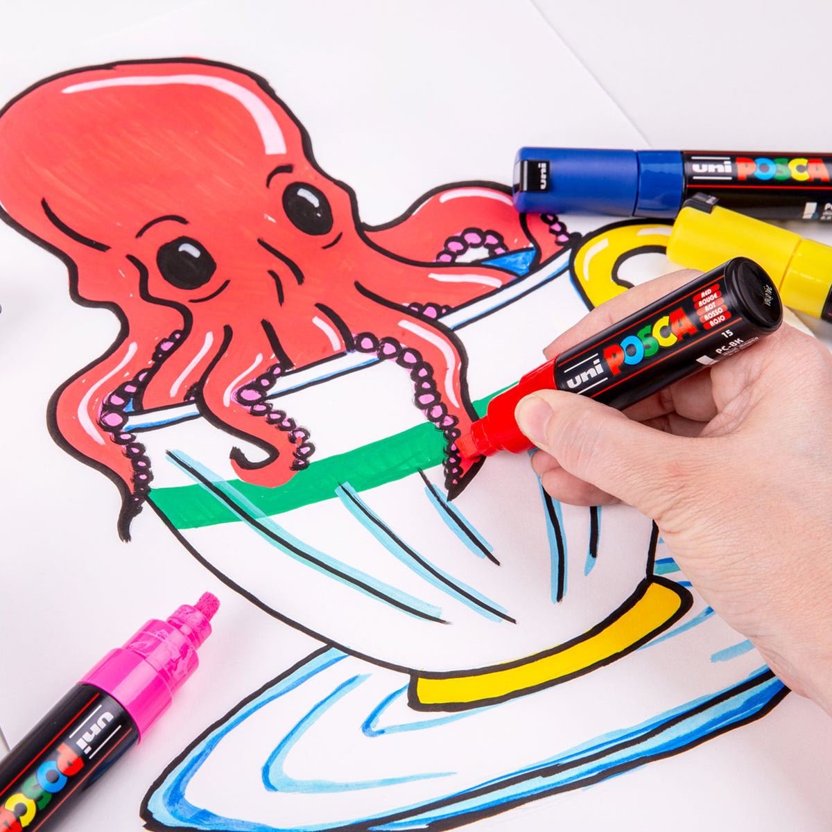 A hand holding a red POSCA Chisel Tip Marker, colouring in a green stripe on a drawn teacup beneath a cartoon red octopus. Blue, yellow and pink capped markers sit nearby on the partly finished illustration.