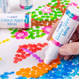 Hand using a Zart Colour Apps Minis bottle to stamp bright orange dots onto white paper. Overlapping circular marks in blue, pink, green and brown form a colourful dot artwork, with additional Colour Apps Minis bottles visible in the background.
