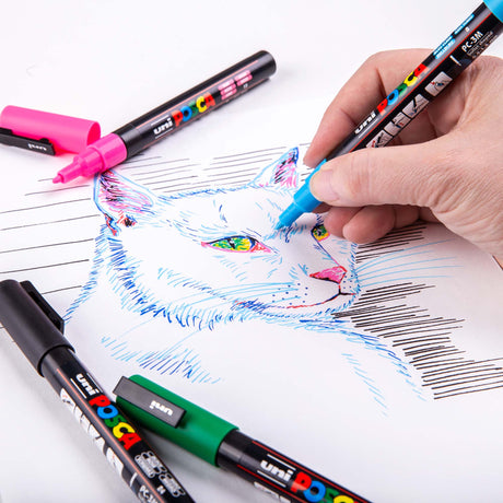 Hand using a blue POSCA marker to draw details on a colourful line-art illustration of a cat. Other POSCA markers lie around the workspace, including a pink uncapped marker.