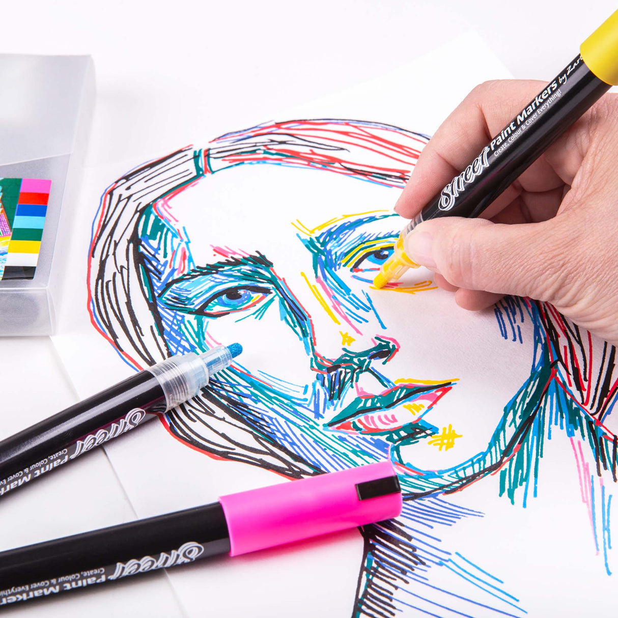 Hand using a yellow Zart Street Paint Marker to add bright strokes to a colourful line-drawn portrait. Other markers and the storage case sit nearby on the workspace.