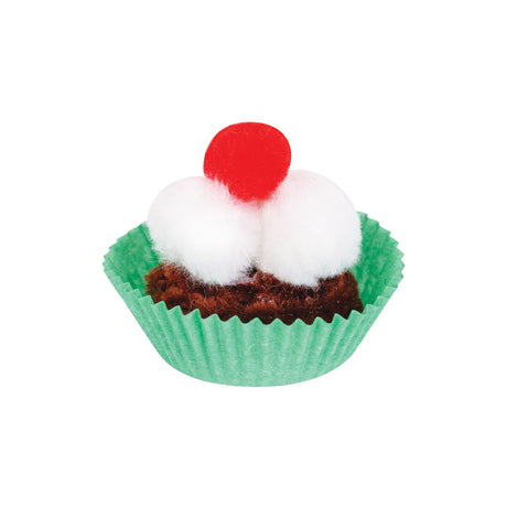 A sample artwork cupcake made using pom poms. Two large white pom poms form the cream topping, with a smaller red pom pom on top as a cherry. Brown pipe cleaners fill the base of a green fluted paper cupcake case.