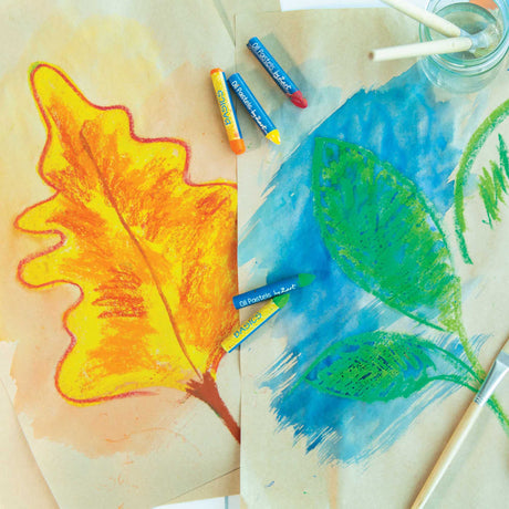 Oil Pastels by Zart Basics sticks scattered across watercolour paintings of autumn leaves in yellow, orange and red, and green leaves on a blue background. The blue-wrapped pastels in yellow, orange, red and green sit alongside a paintbrush and water jar.