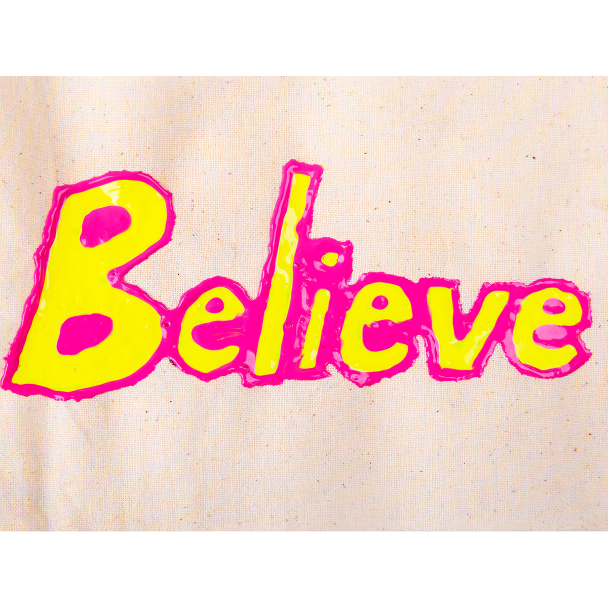 A close-up of the word “Believe” rendered in raised Tulip Slick dimensional fabric paint on natural calico fabric. The bubble lettering is filled in bright yellow with a thick neon pink outline, with glossy pooled texture visible across the surface of the dried paint.