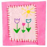 A square of pale pink fabric, hand-stitched with red thread onto a pink felt backing, decorated in raised puffy paint. The design shows a white cloud, a yellow sun with radiating rays, and three flowers in purple, pink and teal with green stems and leaves.