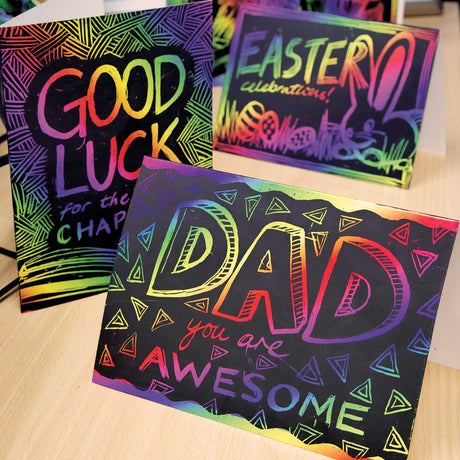 Three Zart Scratch Greeting Cards displayed upright on a wooden table, hand-scratched with rainbow-coloured designs: "GOOD LUCK for the CHAP..." bordered with striped patterning, "EASTER celebrations!" featuring a bunny and eggs, and "DAD you are AWESOME" surrounded by scattered triangle shapes.
