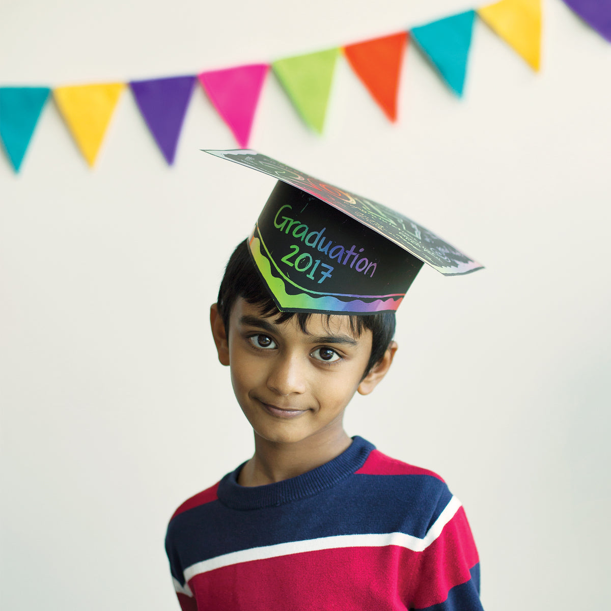 A child wears a decorated Scratch Graduation Hat, its black surface scratched to reveal rainbow-coloured patterns and the words "Graduation 2017" in bright green, blue and purple lettering above a rainbow band. Multicoloured triangular bunting hangs across the white wall behind, and the child smiles at the camera.