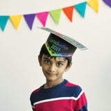 A child wears a decorated Scratch Graduation Hat, its black surface scratched to reveal rainbow-coloured patterns and the words "Graduation 2017" in bright green, blue and purple lettering above a rainbow band. Multicoloured triangular bunting hangs across the white wall behind, and the child smiles at the camera.