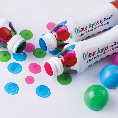 Close-up of Zart Colour Apps bottles lying on their sides with exposed sponge tips. Bright blue, pink and green circular paint marks are stamped onto white paper, with loose caps nearby showing the rounded applicator tops.