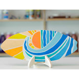 Painted surfboard artwork displayed horizontally on a wooden stand. The design features abstract waves and sun shapes in blue, teal, yellow and orange, with a softly blurred studio background.