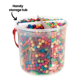 A clear plastic storage tub with a red carry handle, filled with Zart Basics plastic beads in assorted shapes and bright colours, divided into sections by card inserts. The annotation "Handy storage tub" with an arrow points to the container from the upper left.
