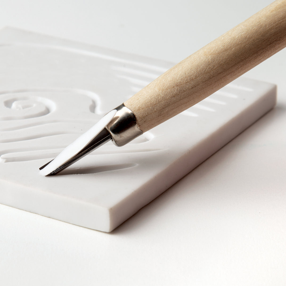 Close-up of a wooden-handled carving tool cutting into a white soft cut block. The metal blade forms shallow recessed lines within a curved design. The block surface appears smooth, with clean edges and visible carved grooves.