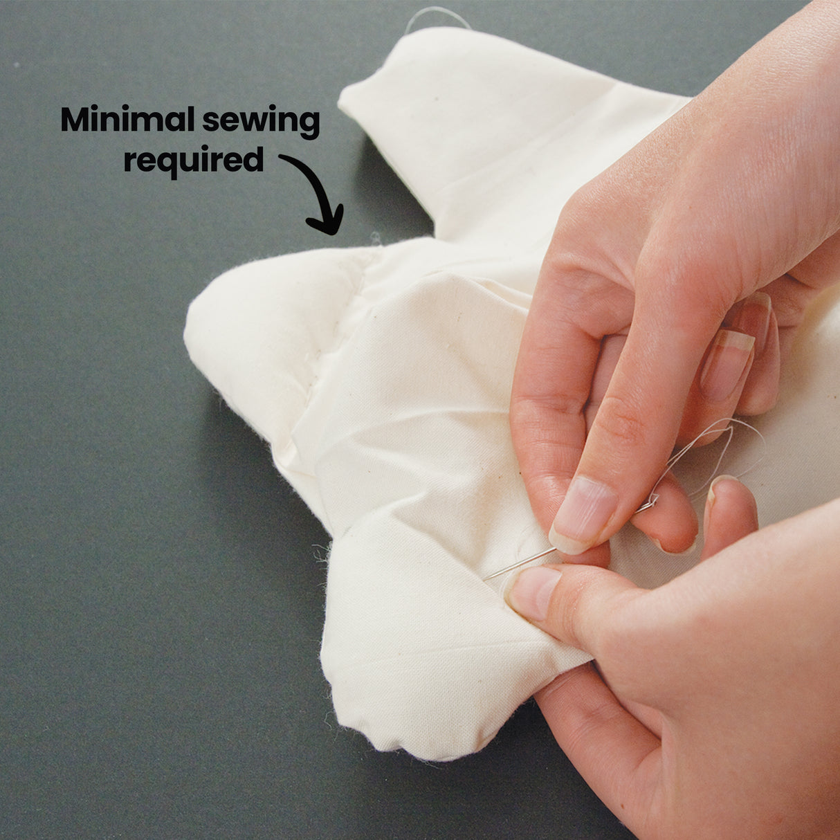 A close-up of hands hand-stitching the opening of a cream calico bear closed with a needle and thread. A bold black annotation reads "Minimal sewing required" with an arrow pointing to the stitching area. Shown on a dark grey surface.