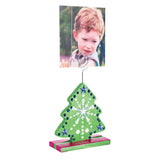 A decorated Zart Photo Holder Tree painted green with navy and white beads, silver stars, and a snowflake motif, set on a pink and green striped base. The spiral wire clip holds a photograph of a young boy on a swing above the tree shape.